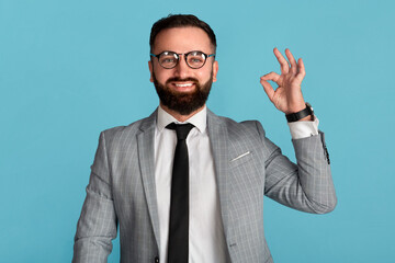 Smiling businessman in formal suit showing okay gesture on blue background