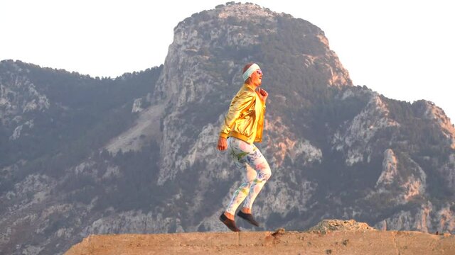 Young bald slender freak man caucasian in tight leggings, gold jacket, bandage on his head, dancing on roof at sunset, runs, has fun, behaves unusually. Slow motion video. Fun, humor, unusual people