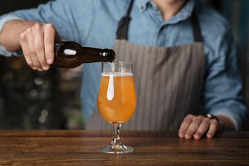 Ale for company. Barman in apron pours light beer from bottle in glass on wooden bar counter