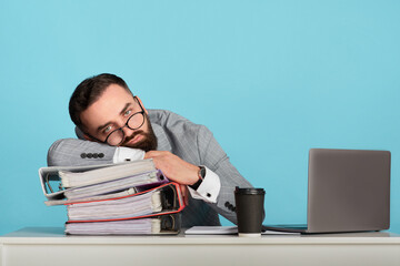 Overworked businessman lying on desk near laptop computer against blue background, blank space