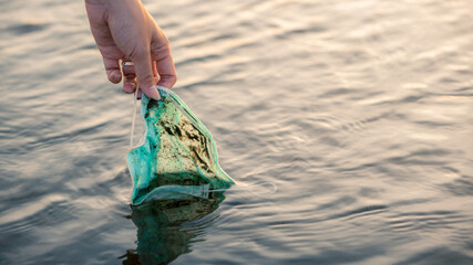 Woman hand picking up discarded used disposable medical mask in sea waters