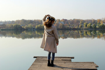Young girl wearing a beige trench coat, jeans and black boots, standing on a wooden bridge and looking to the beautiful landscape. Young girl standing near the lake. People, nature, travel concept.