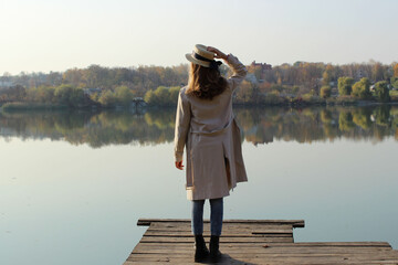 Young girl wearing a beige trench coat, jeans and black boots, standing on a wooden bridge and looking to the beautiful landscape. Young girl standing near the lake. People, nature, travel concept.
