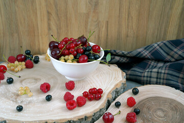  Raspberries, cherries, white and red currants, black currants in a white bowl on wooden pieces of tree trunk