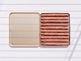 Cigarillos are in an open tin box. A shadow from the blinds falls on a white background. Tobacco smoking concept.