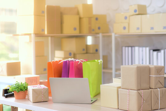 On The Table There Are Laptops, Shopping Bags And Parcel Boxes, Background Is A Shelf Full Of Brown Boxes. Of Small Businesses By Selling Products Online, Work From Home And Online Shopping Concept