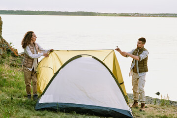 Contemporary young hikers putting tent on the ground by waterside before night