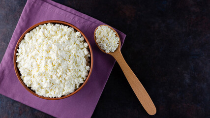 Cottage cheese and wooden spoon on a dark background. Fresh cottage cheese in a bowl. Clay plate with soft cheese on a linen napkin. Top view. Copy space