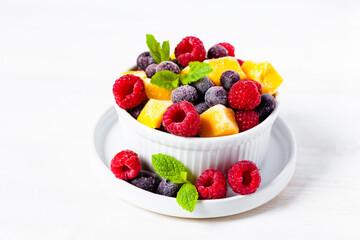 Frozen fruits: mango, raspberry, blueberry with fresh green mint in white bowl. Concept of healthy eating, delicious low calories dessert, summer refreshing light meal. White background copy space