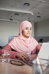 Vertical portrait of young businesswoman wearing headscarf using laptop at desk while working in office alone, copy space