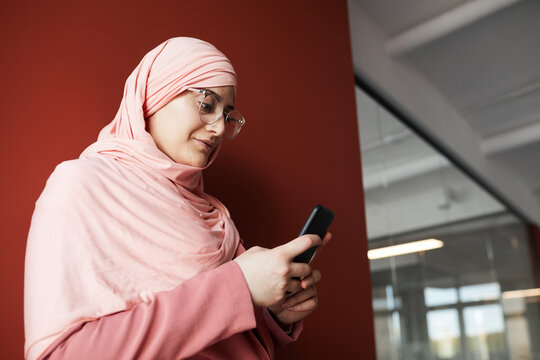Side view portrait of young Muslim businesswoman using smartphone while standing against red wall in office, copy space