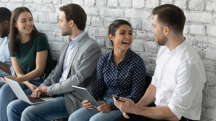 Cheerful diverse applicants hold devices talking while waiting turn job interview. People gather in office prepared for seminar, communicating enjoy chat and friendly relations in collective concept
