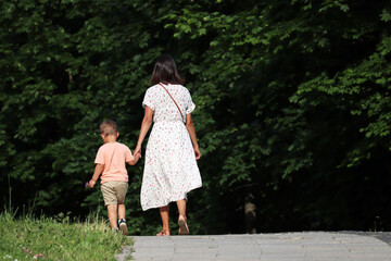 Mother and kid are walking down the path holding hands. Young woman in dress lead little boy in a summer park, concept for motherhood, single mother or babysitter