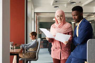 Waist up portrait of modern Muslim businesswoman talking to African-American colleague and...
