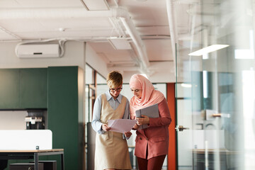 Wide angle portrait of female manager talking to young Muslim businesswoman and holding documents while walking towards camera in office interior, copy space