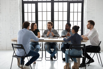 Six multi racial young and skilled employees gathered in boardroom sit at desk share ideas involved...