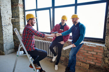 Fototapeta premium Three young architects in protective helmets clinking with glasses of coffee