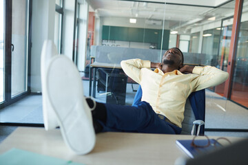 Portrait of contemporary African-American businessman relaxing at workplace in office with feet on desk, copy space