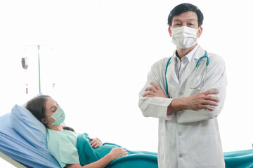 Portrait of doctor wearing a mask with stethoscope on white background, confident medical doctor standing and crossing arm with patient lying on the hospital bed behind him, medical health care and ho