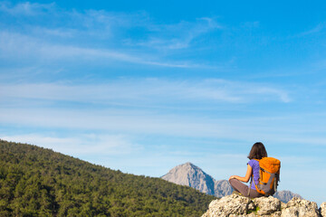 Fototapeta na wymiar A woman with a backpack sits on top of a mountain and admires the beauty of a mountain valley.