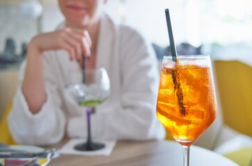 Glass of cold refreshing aperol spritz cocktail on table in summer. Woman in bathrobe in background. 