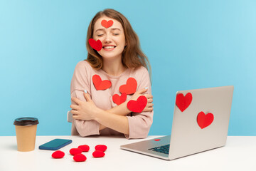 Charming happy young woman with kind smile sitting at workplace office, all covered with sticker love hearts, embracing herself, enjoying valentine's day greetings. indoor studio shot, isolated © khosrork