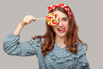 Playful pinup girl ruffle blouse covering eye with sweet spiral candy looking at camera, showing tongue out, having fun with lollipop. retro vintage style. indoor studio shot isolated gray background,