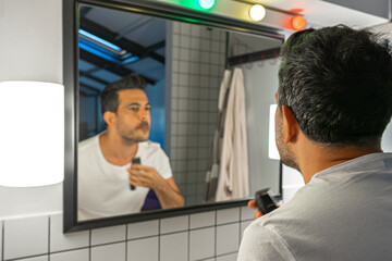 handsome man is shaving his beard with trimmer machine in front of bathroom mirror