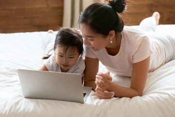 Happy small cute asian baby girl lying on bed with smiling caring mother or babysitter, learning...