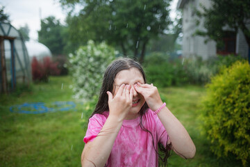 portrait of happy wet child in rain in garden in hot summer. kids fun outroors on vacation. rainy weather. girl wipes her wet eyes with hands