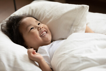 Close up smiling adorable little asian ethnic kid girl lying under warm blanket on pillow, feeling energetic after waking up at holiday morning, joyful small biracial baby having fun alone in bed.