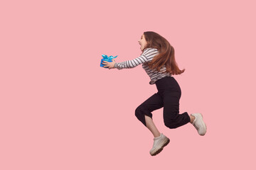 Full length side view, woman running quickly with gift box, hurrying and shouting for joy, expressing great excitement, flying with birthday present. indoor studio shot isolated on pink background