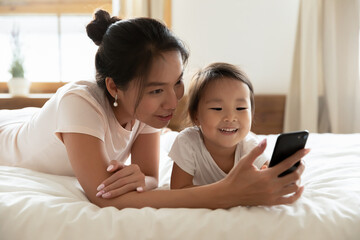 Smiling young asian ethnic woman lying on cozy bed, showing mobile application, video game or online cartoons to funny cute little biracial adopted baby daughter, posing for family selfie in bedroom.