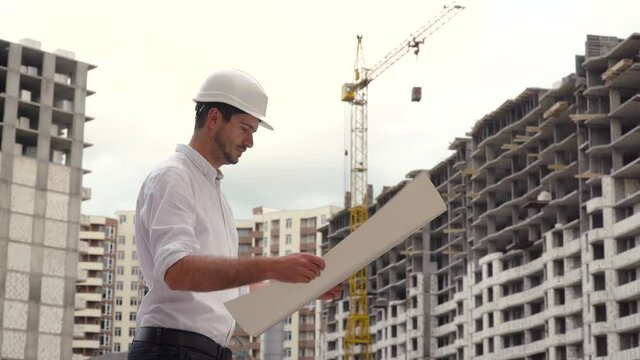 Portrait of a successful young engineer, architect, builder, businessman, wearing a white helmet, in a shirt, holding a project in his hand, a skyscraper background and a construction site