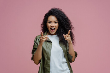 Glad confident woman with Afro hair points index fingers directly at camera, chooses you, picks someone in her team, dressed in white t shirt and leather shirt, poses against pink background