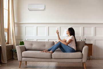 Full length smiling young asian ethnic woman sitting on cozy sofa, turning on off air conditioner...