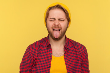 Portrait of hipster guy in beanie hat and checkered shirt making disobedient face showing tongue...