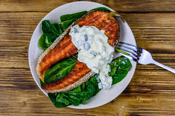 Plate with roasted salmon steak, tartar sauce and spinach leaves on a wooden table. Top view