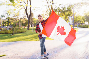 A young guy of 9 years old riding on a sunny road in the evening in the city park, dressed in a flannel plaid shirt. Holding fluttering flag of Canada. Happy Day Independence day to Canada