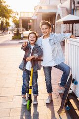 two attractive teenagers of Caucasian nationality 7 and 9 years old standing on sunny evening street in the city. Laughing, pointing fingers forward, holding scooter and skateboard