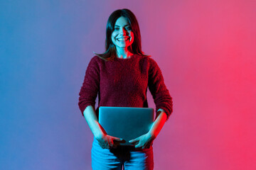 Portrait of successful happy office worker, pretty girl in casual pullover standing, holding closed laptop or folder, looking at camera with toothy smile, positive expression. neon light, studio shot © khosrork