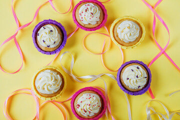 Six multi-colored cupcakes with cream on a yellow background with different ribbons in the form of a circle, top view. Festive background. Holiday baking.
