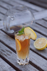 A glass of ice tea with lemon and mint, on a rustic gray aged table with a glass tea pot in the background and half a lemon