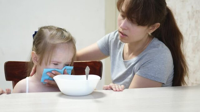 Mom Calms A Capricious Child Sitting At The Table. Parent Wipes Kid Drool And Snot With A Napkin. Teamwork. Baby Doesn T Want To Eat Porridge