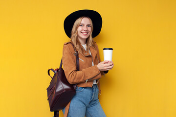 girl tourist in autumn clothes with a backpack and coffee smiling on a yellow isolated background, a woman warmly dressed in autumn, copy space