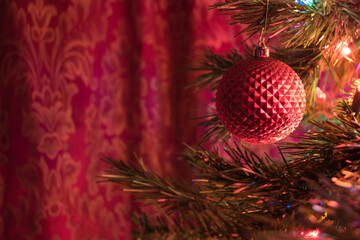 Red Christmas ornament against a red background with glowing holiday lights against an evergreen tree.