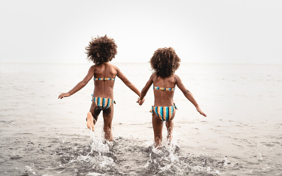 Happy sisters running inside water during summer time - Afro kids having fun playing on the beach - Family love and travel vacation lifestyle concept