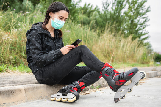 Woman in protective face mask, on roller skating pause, sitting on the street and using mobile phone during coronavirus pandemic outbreak. Urban Girl using phone, wearing Roller Skates .