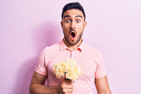 Young handsome romantic man with beard holding bouquet of flowers over pink background scared and amazed with open mouth for surprise, disbelief face