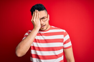 Young handsome man wearing casual striped t-shirt and glasses over isolated red background Yawning tired covering half face, eye and mouth with hand. Face hurts in pain.